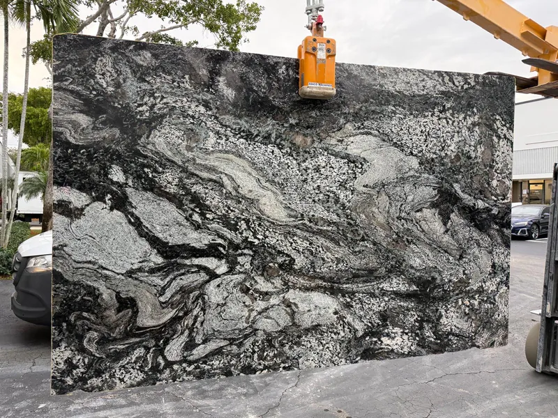 A full stone slab with a bold black and white swirl, held up by a vacuum lifter in a parking lot