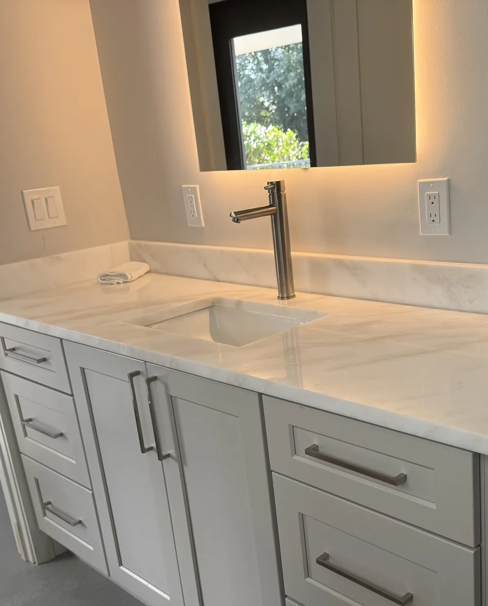 White vanity top with soft gray veining, an undermount sink, a tall single-hole faucet and a matching backsplash below a backlit mirror, over gray shaker drawers