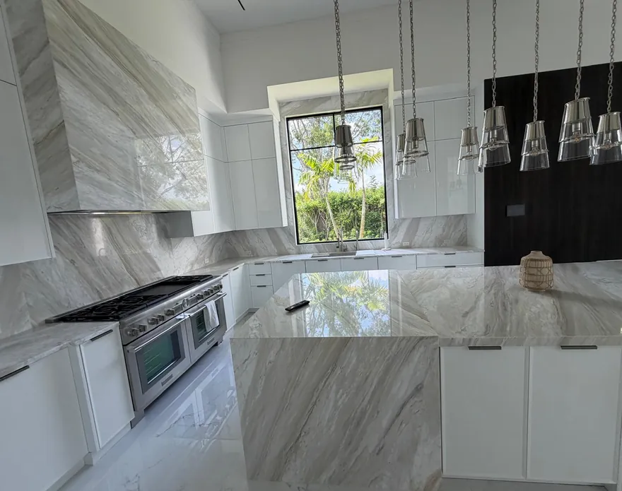 Kitchen with a waterfall island, a stone-clad range hood and a matching full-height backsplash under glass pendant lights