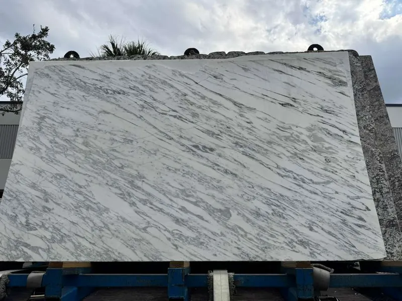 Full stone slab standing on a blue A-frame rack outdoors, gray veining running corner to corner across a white face