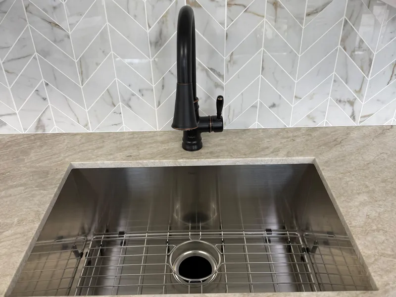 Stainless steel undermount sink with a bottom grid under a veined countertop, below a dark bronze pull-down faucet and a chevron tile backsplash