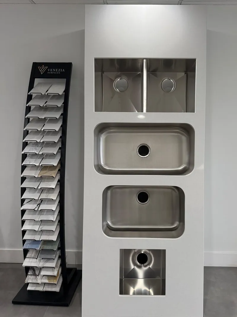 Showroom display panel with four stainless steel sink bowls cut into one white slab, next to a rack of surface samples