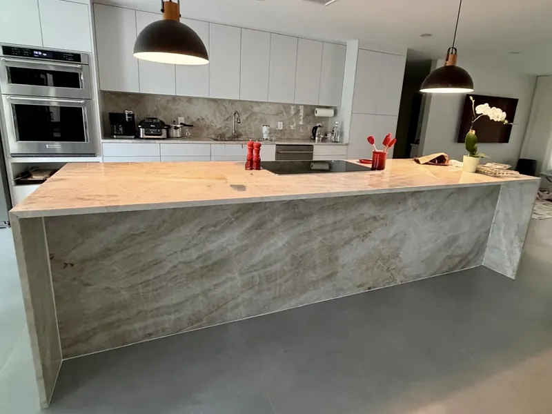 Taj Mahal quartzite waterfall island with a cooktop, white flat-panel cabinets and two black pendant lights